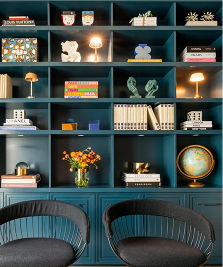 Estantería azul oscuro diseñada por Mónica Priale, decorada con libros, esculturas, lámparas y objetos artísticos; en la parte inferior se observan dos sillas modernas y un florero con flores naranjas.